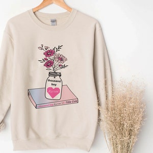 May include: A beige sweatshirt with a graphic of a mason jar filled with pink flowers and a heart that says "kisses". The jar is sitting on top of a book that says "A Thousand Boy Kisses" by Tillie Cole.