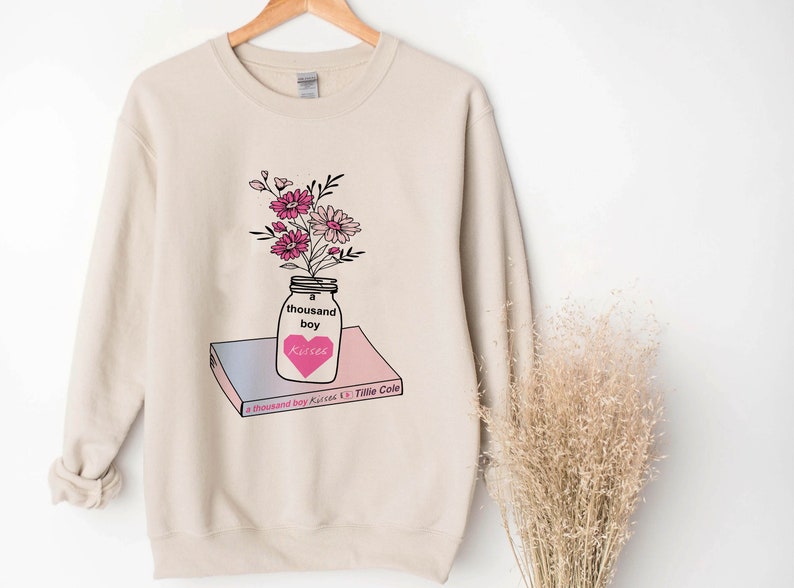 May include: A beige sweatshirt with a graphic of a mason jar filled with pink flowers and a heart that says "kisses". The jar is sitting on top of a book that says "A Thousand Boy Kisses" by Tillie Cole.