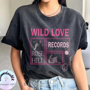 May include: A black t-shirt with a vintage aesthetic featuring the text "WILD LOVE RECORDS" in pink lettering. The design also includes a rose, a hand holding a rose, a stack of books, and a key with a heart.