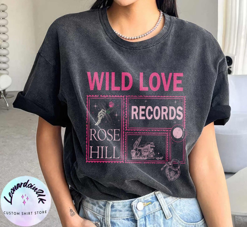 May include: A black t-shirt with a vintage aesthetic featuring the text "WILD LOVE RECORDS" in pink lettering. The design also includes a rose, a hand holding a rose, a stack of books, and a key with a heart.