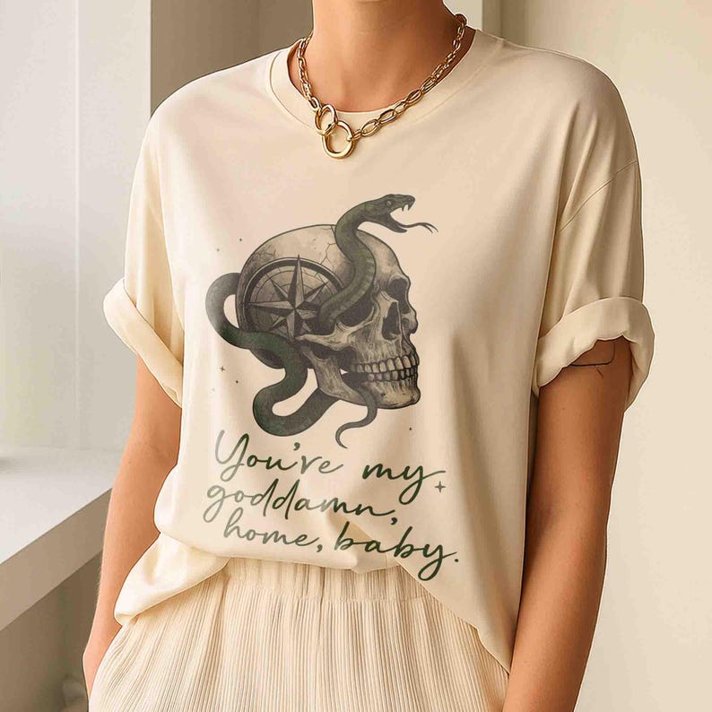 May include: Cream-colored t-shirt featuring a skull with a compass design and a green snake wrapped around it. The shirt has the text "You're my goddamn home, baby" in a handwritten style.