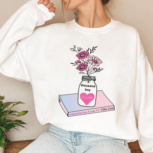 May include: White sweatshirt with a graphic of a mason jar filled with pink and white flowers. The jar is sitting on a book with the title "a thousand boy Kisses" by Tillie Cole.