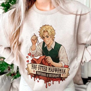 May include: Beige sweatshirt featuring a cartoon illustration of a blond-haired person in a green vest, sewing red fabric. The text reads "YOU UTTER MADWOMAN, PROFESSOR OF DRYADOLOGY" on a banner with heart accents.