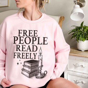 May include: A light pink sweatshirt with the text "FREE PEOPLE READ FREELY" in black. Below the text is a graphic of a stack of books and a lit candle. The sweatshirt is long-sleeved and has a crew neck.