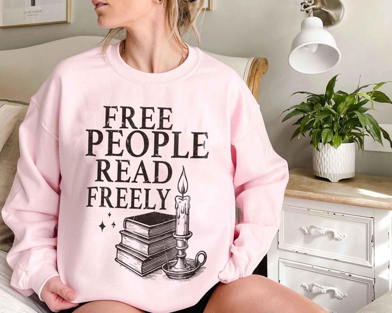 May include: A light pink sweatshirt with the text "FREE PEOPLE READ FREELY" in black. Below the text is a graphic of a stack of books and a lit candle. The sweatshirt is long-sleeved and has a crew neck.