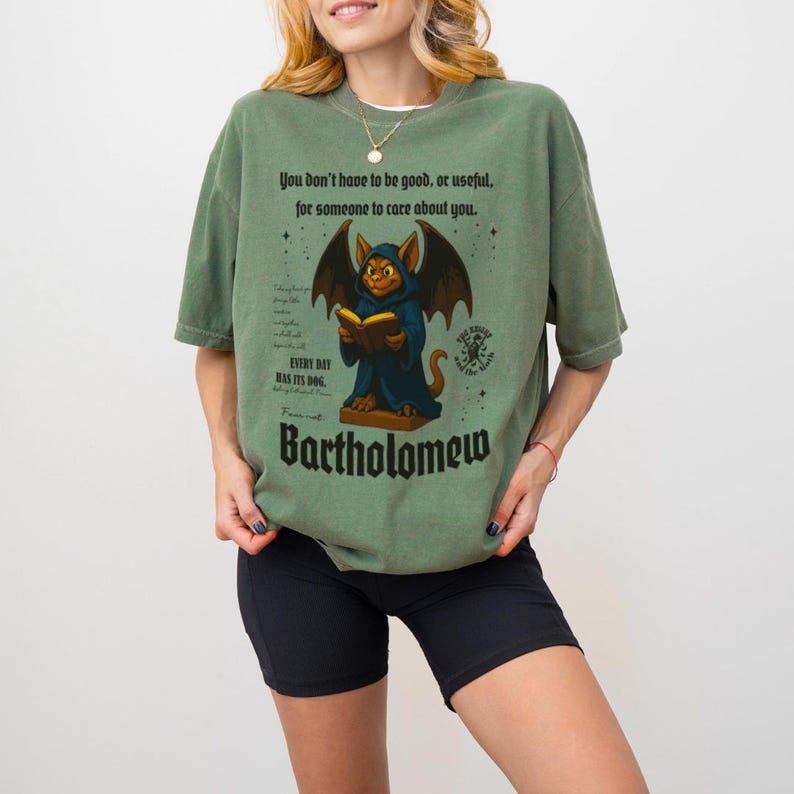 May include: Sage green t-shirt featuring a cartoon bat-like creature in a hooded robe, reading a book. The shirt includes the text "You don't have to be good, or useful, for someone to care about you." and "Bartholomew."