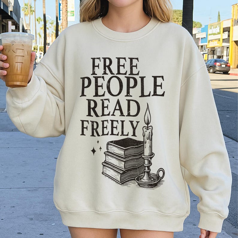 May include: Cream-colored sweatshirt with the text "FREE PEOPLE READ FREELY" in black. Below the text is a graphic of stacked books, a candle, and two small stars. The sweatshirt is a casual style.