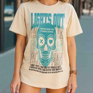 Pu&ograve; includere: T-shirt color crema con grafica a teschio turchese e la scritta "LIGHTS OUT". Il teschio ha una X e un cuore al posto degli occhi. Il testo aggiuntivo include "I NEED MORE THERAPY THAN I'M CURRENTLY RECEIVING" e una citazione.