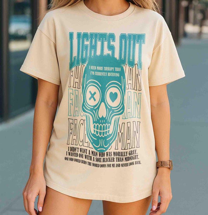Pu&ograve; includere: T-shirt color crema con grafica a teschio turchese e la scritta "LIGHTS OUT". Il teschio ha una X e un cuore al posto degli occhi. Il testo aggiuntivo include "I NEED MORE THERAPY THAN I'M CURRENTLY RECEIVING" e una citazione.