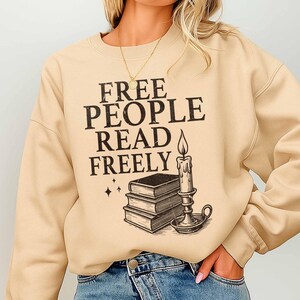 May include: Beige sweatshirt with the text "FREE PEOPLE READ FREELY" in black, along with a graphic of stacked books, a candle, and stars. The sweatshirt has a crew neck and long sleeves.