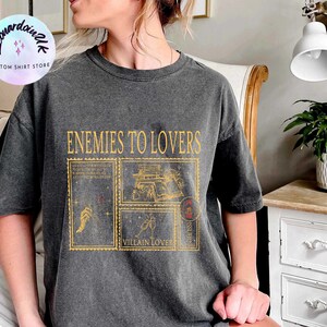 Puede incluir: Una camiseta gris oscuro con un estampado gr&aacute;fico de estilo vintage que dice "Enemies to Lovers" con un dise&ntilde;o de cuatro sellos postales con ilustraciones de un libro, una mano con una varita, una copa de champ&aacute;n y un coraz&oacute;n con una flecha.