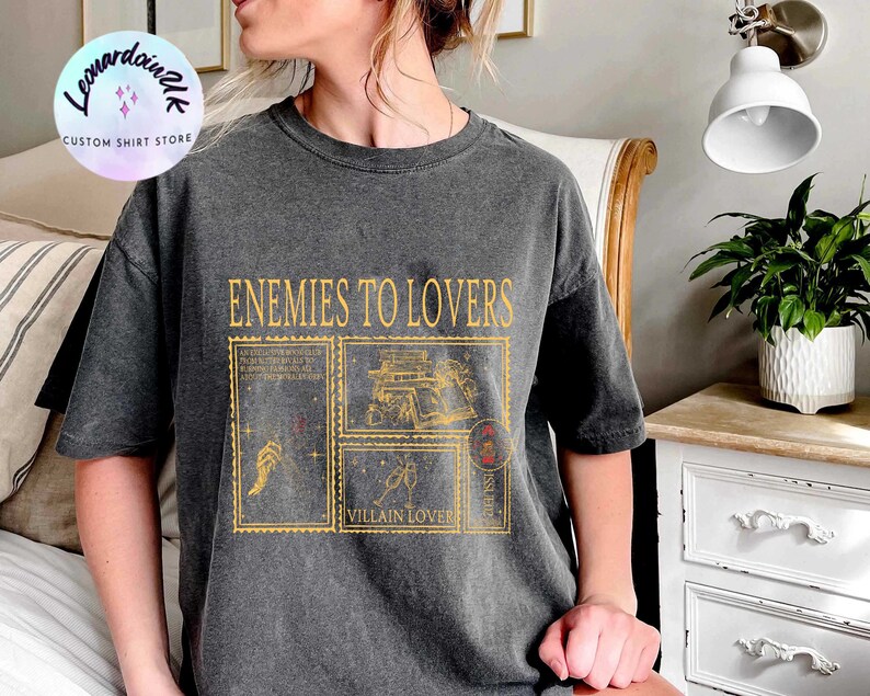 Puede incluir: Una camiseta gris oscuro con un estampado gr&aacute;fico de estilo vintage que dice "Enemies to Lovers" con un dise&ntilde;o de cuatro sellos postales con ilustraciones de un libro, una mano con una varita, una copa de champ&aacute;n y un coraz&oacute;n con una flecha.