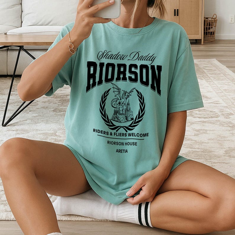 May include: A light teal t-shirt featuring the text "Shadow Daddy RIORSON" in black, above a circular emblem with a dragon and castle design. Below the emblem are the words "RIDERS & FLIERS WELCOME RIORSON HOUSE ARETIA".
