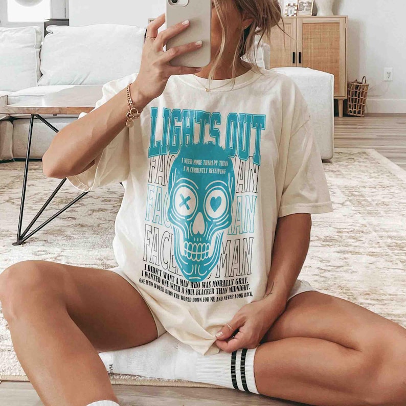 Pu&ograve; includere: T-shirt color crema con grafica a teschio turchese e la scritta "LIGHTS OUT FACE MAN." Il testo aggiuntivo include "I need more therapy than I'm currently receiving." e una citazione. La maglietta &egrave; in stile casual e oversize.
