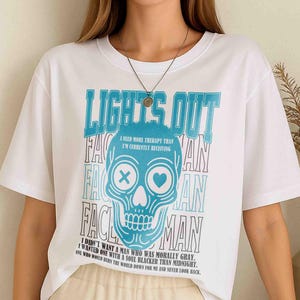 Pu&ograve; includere: T-shirt bianca con grafica di teschio blu e la scritta "LIGHT'S OUT". Il teschio ha una X per un occhio e un cuore per l'altro. Il testo aggiuntivo include "I NEED MORE THERAPY THAN I'M CURRENTLY RECEIVING".