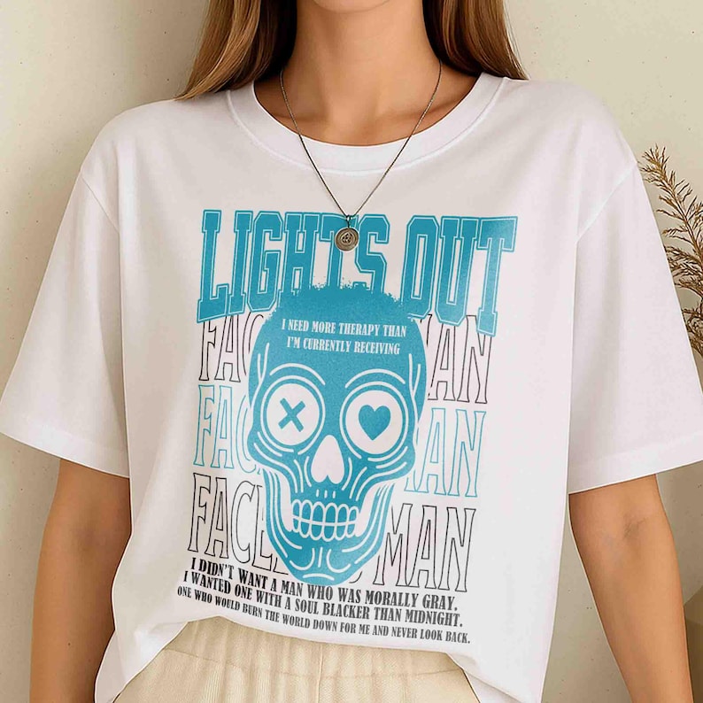 Pu&ograve; includere: T-shirt bianca con grafica di teschio blu e la scritta "LIGHT'S OUT". Il teschio ha una X per un occhio e un cuore per l'altro. Il testo aggiuntivo include "I NEED MORE THERAPY THAN I'M CURRENTLY RECEIVING".