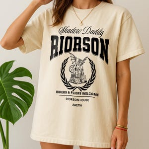 May include: A cream-coloured t-shirt with the text "Shadow Daddy RIORSON" in black, above a circular emblem featuring a dragon and castle design. The shirt also includes the text "RIDERS & FLIERS WELCOME" and "RIORSON HOUSE ARETIA".