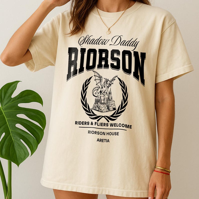 May include: A cream-coloured t-shirt with the text "Shadow Daddy RIORSON" in black, above a circular emblem featuring a dragon and castle design. The shirt also includes the text "RIDERS & FLIERS WELCOME" and "RIORSON HOUSE ARETIA".