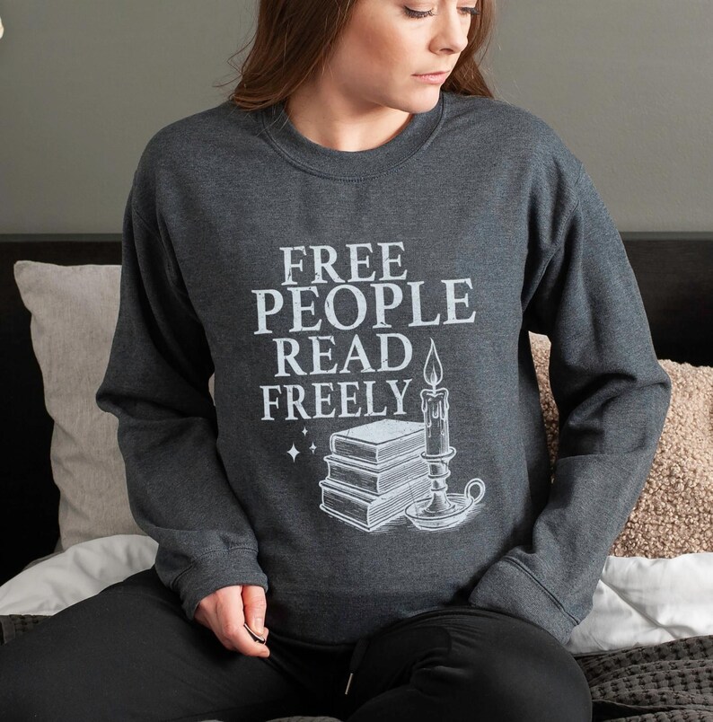 May include: Dark gray sweatshirt with the text "FREE PEOPLE READ FREELY" in white. The graphic includes a stack of books, a candle, and stars. The sweatshirt is long-sleeved and has a crew neck.