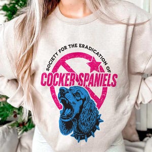 May include: Beige sweatshirt with a graphic that reads "SOCIETY FOR THE ERADICATION OF COCKER SPANIELS." The graphic features a blue Cocker Spaniel with an open mouth inside a pink circle with a star.