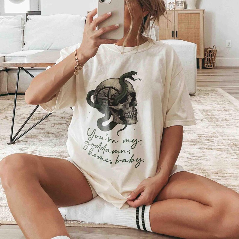 May include: Cream-colored t-shirt featuring a skull with a compass design and a green snake. The shirt has the text "You're my goddamn home, baby" in a script font. The shirt is a casual, relaxed fit.