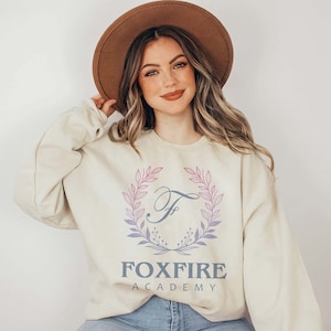 May include: A cream-colored sweatshirt with a pink and purple floral wreath design and the text "FOXFIRE ACADEMY" in gray.