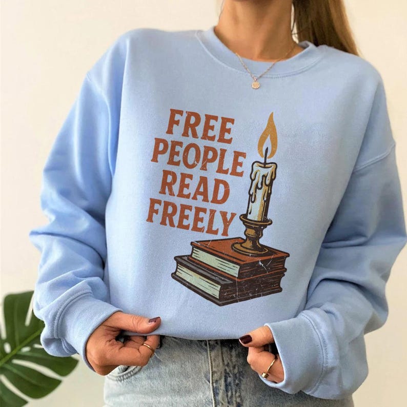 May include: A pale blue sweatshirt featuring the text "FREE PEOPLE READ FREELY" in brown. A candle with a lit flame is on a stack of books. The sweatshirt has long sleeves.