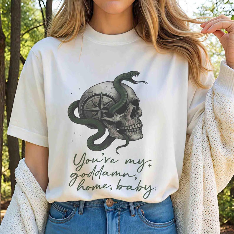 May include: Off-white t-shirt featuring a skull with a compass design and a green snake wrapped around it. The text "You're my goddamn home, baby" is printed in green script below the graphic.