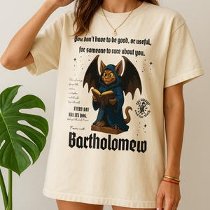 May include: Cream-colored t-shirt featuring a cartoon gargoyle reading a book, with the name "Bartholomew" below. The shirt includes the text "You don't have to be good, or useful, for someone to care about you."