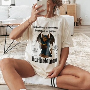 May include: Cream-colored t-shirt featuring a graphic of a gargoyle reading a book, with the name "Bartholomew" below. The shirt includes the text "You don't have to be good, or useful, for someone to care about you."