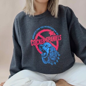 May include: Dark gray sweatshirt with a graphic featuring a blue Cocker Spaniel inside a red circle with the text "SOCIETY FOR THE ERADICATION OF COCKER SPANIELS" in red and blue.