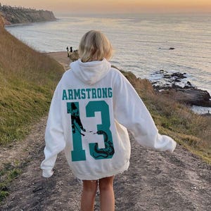 May include: A white hoodie with the name "ARMSTRONG" and the number "13" in teal. The number "13" features a graphic of a hockey player and a snake. The hoodie is worn outdoors with a scenic ocean view in the background.