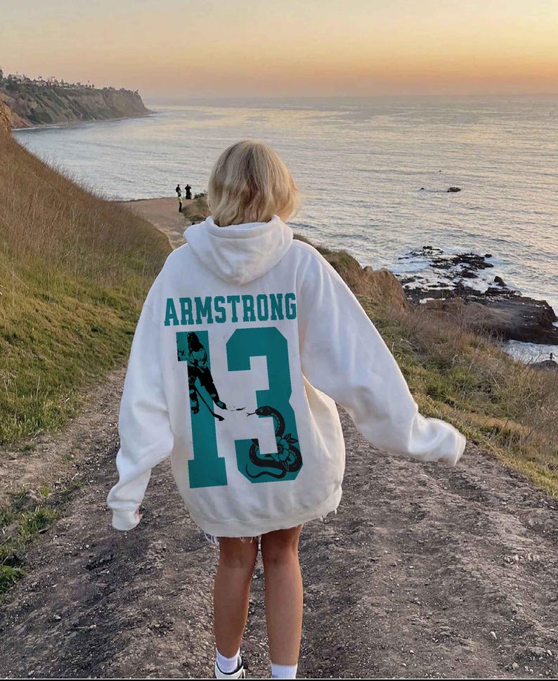 May include: A white hoodie with the name "ARMSTRONG" and the number "13" in teal. The number "13" features a graphic of a hockey player and a snake. The hoodie is worn outdoors with a scenic ocean view in the background.