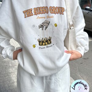 May include: White sweatshirt with a graphic print of a hand holding a crown with the text "The Russo Group Luxury Goods Dante Russo" printed on the front.