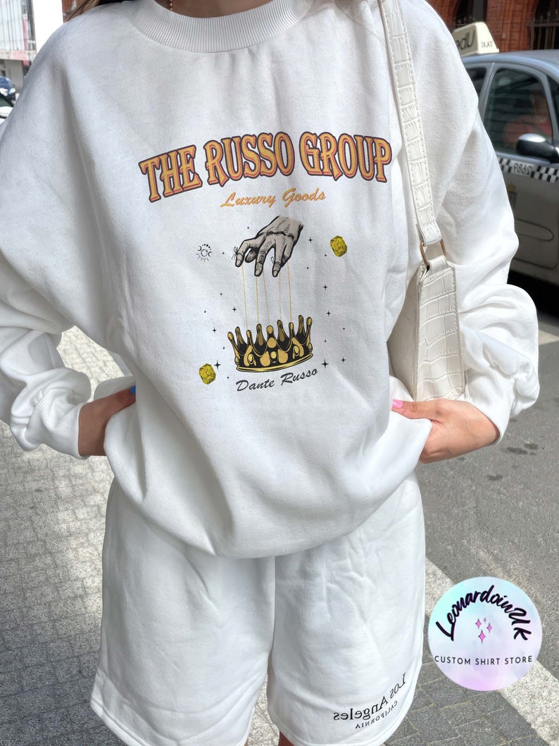 May include: White sweatshirt with a graphic print of a hand holding a crown with the text "The Russo Group Luxury Goods Dante Russo" printed on the front.