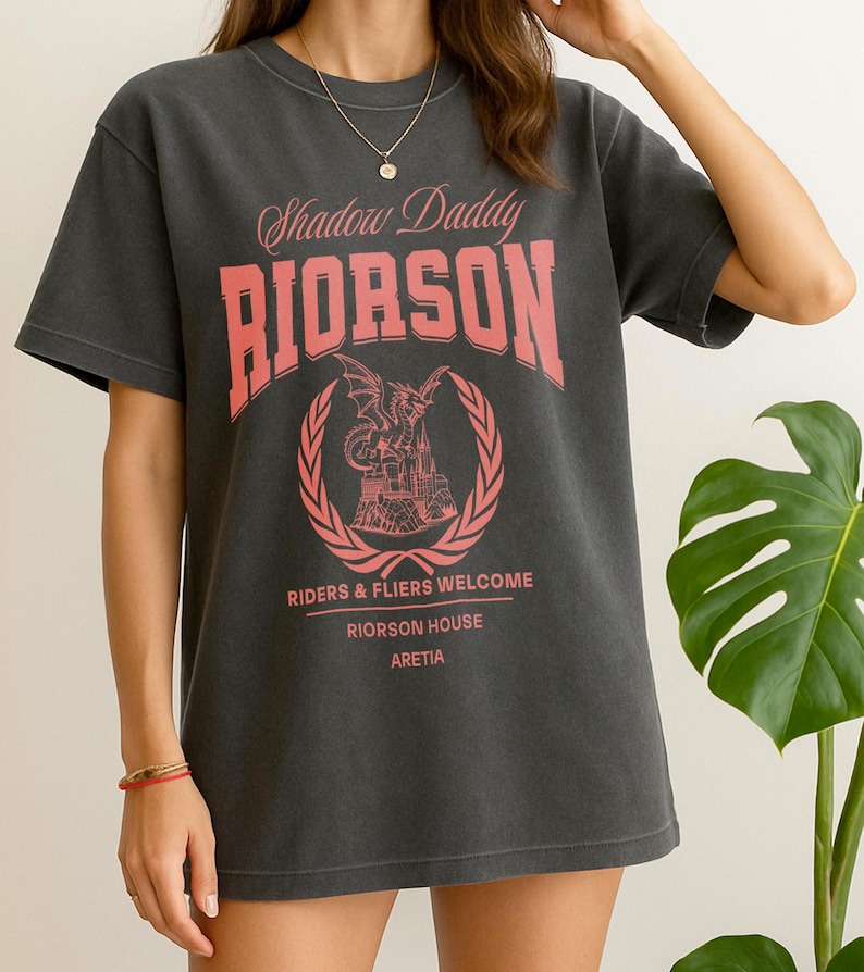 May include: Charcoal grey t-shirt featuring a pink graphic. The design includes the text "Shadow Daddy RIORSON" above a crest with a dragon and castle, and the text "RIDERS & FLIERS WELCOME RIORSON HOUSE ARETIA" below.