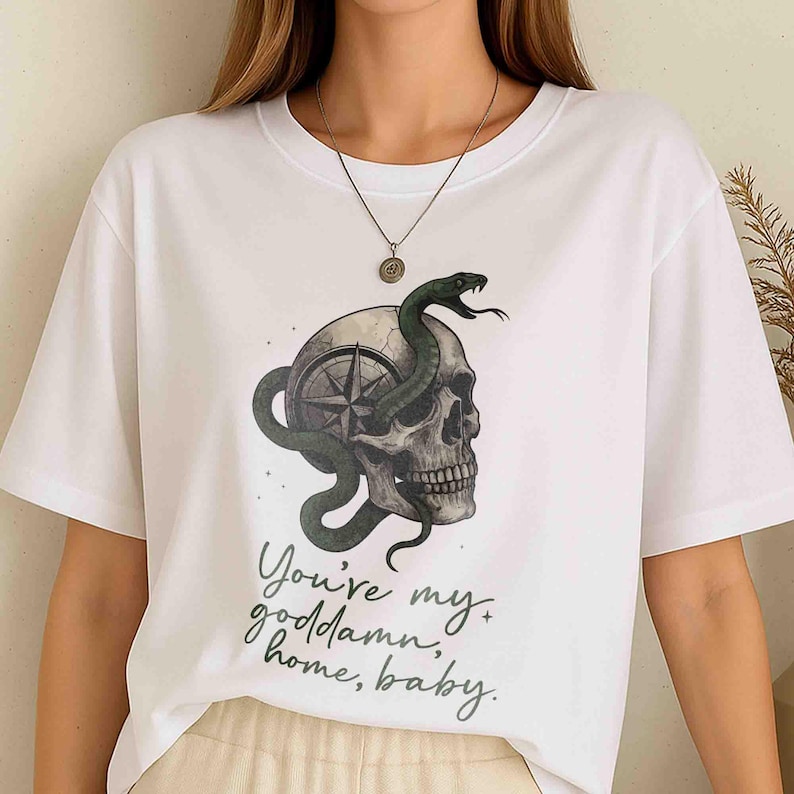 May include: White t-shirt featuring a graphic of a skull with a compass design, entwined by a green snake. The shirt includes the text "You're my goddamn home, baby." A casual, unisex tee.