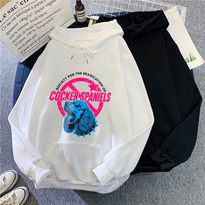 May include: White hoodie with a front pocket and drawstring. The graphic on the front features a blue Cocker Spaniel inside a pink circle with the text "SOCIETY FOR THE ERADICATION OF COCKER SPANIELS". A black hoodie is next to it.