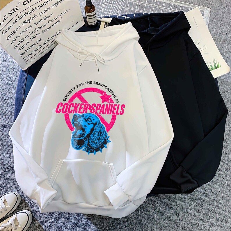 May include: White hoodie with a front pocket and drawstring. The graphic on the front features a blue Cocker Spaniel inside a pink circle with the text "SOCIETY FOR THE ERADICATION OF COCKER SPANIELS". A black hoodie is next to it.