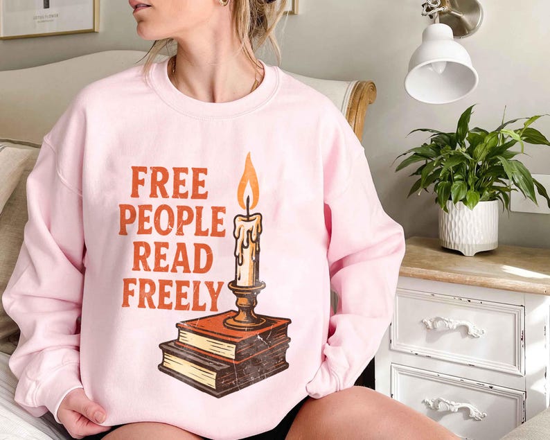 May include: A pale pink sweatshirt featuring the slogan "FREE PEOPLE READ FREELY" in orange and brown. A candle graphic with an orange flame sits above two books. The sweatshirt is a relaxed, casual style.