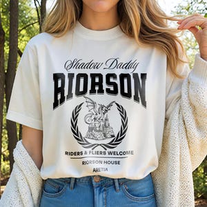 May include: Off-white t-shirt featuring the words "Shadow Daddy RIORSON" in black, with a dragon and castle graphic inside a laurel wreath. The text "RIDERS & FLIERS WELCOME, RIORSON HOUSE, ARETIA" is below.