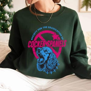 May include: Dark green sweatshirt with a graphic that reads "Society for the Eradication of Cocker Spaniels." The graphic features a blue Cocker Spaniel with an open mouth inside a pink circle with a red slash through it.
