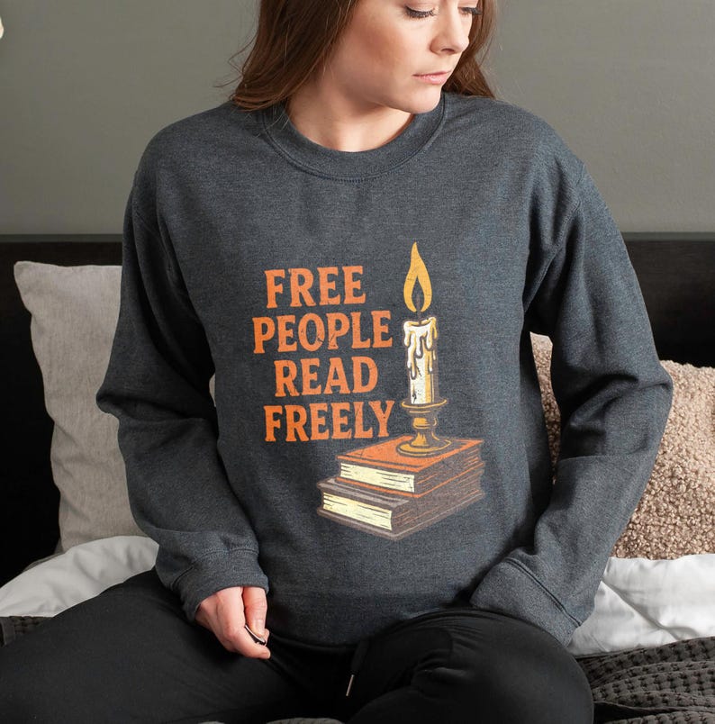 May include: Dark grey sweatshirt featuring the orange text "FREE PEOPLE READ FREELY". A candle with a lit flame is depicted above a stack of books. The sweatshirt has a crew neck design.