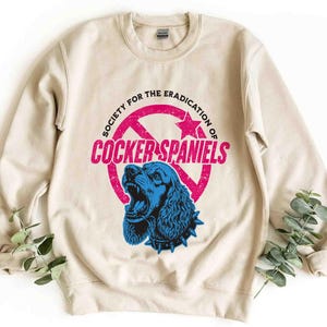 May include: A beige sweatshirt with a graphic that reads "Society for the Eradication of Cocker Spaniels." The graphic features a blue Cocker Spaniel with an open mouth inside a pink circle with a star and arrow design.