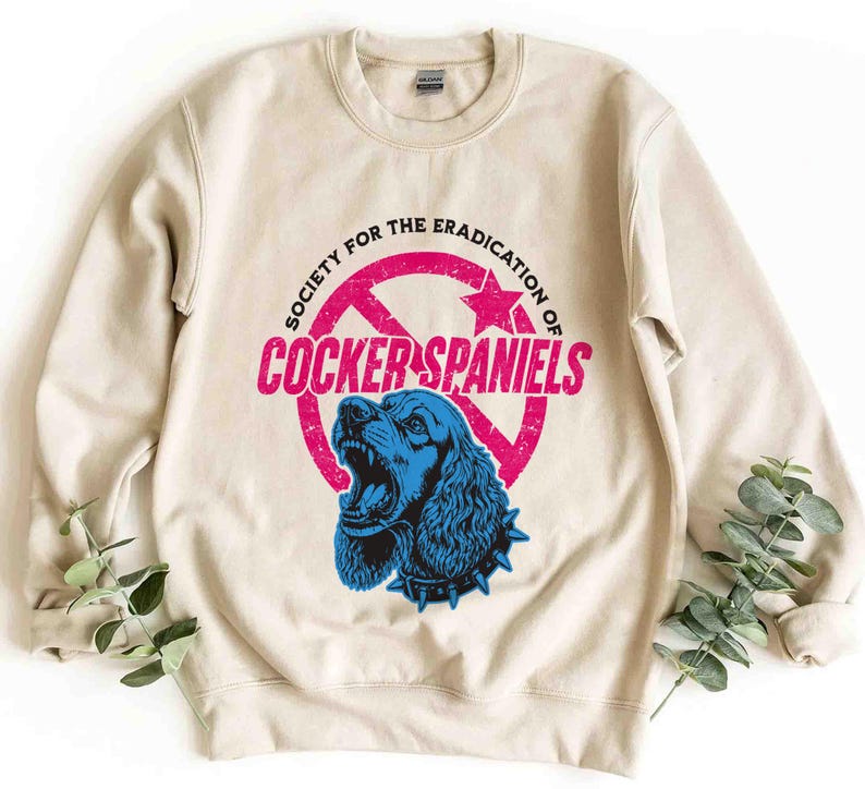 May include: A beige sweatshirt with a graphic that reads "Society for the Eradication of Cocker Spaniels." The graphic features a blue Cocker Spaniel with an open mouth inside a pink circle with a star and arrow design.