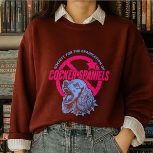 May include: A maroon sweatshirt with a graphic that reads "SOCIETY FOR THE ERADICATION OF COCKER SPANIELS". The graphic features a blue Cocker Spaniel head inside a pink circle with a star.