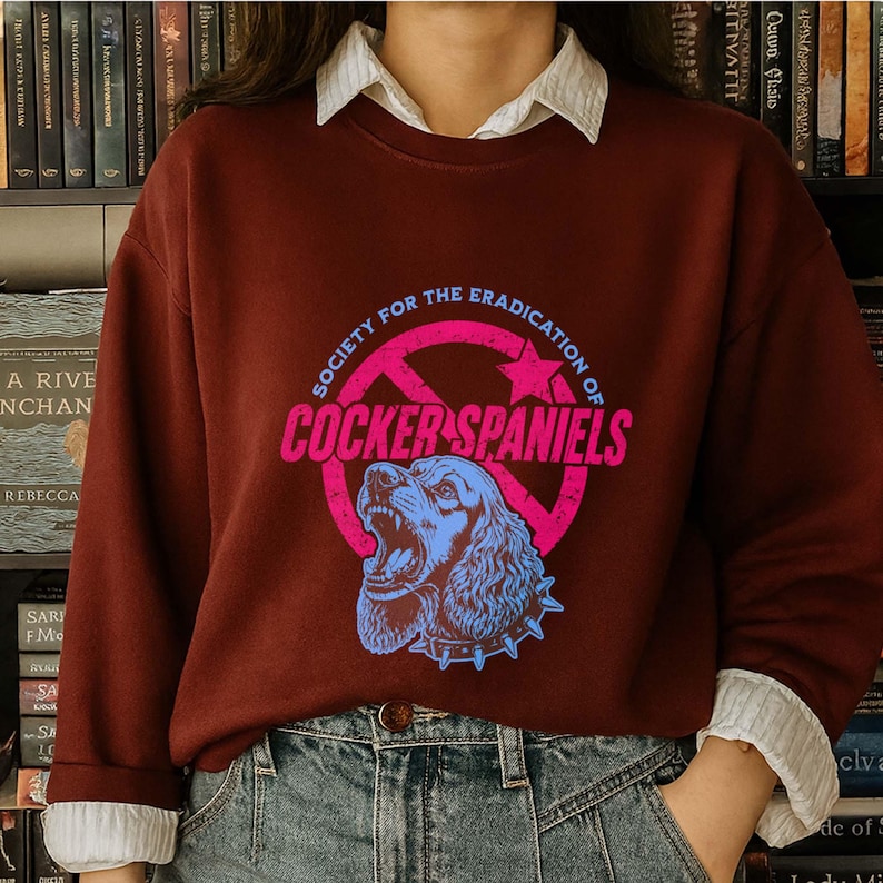 May include: A maroon sweatshirt with a graphic that reads "SOCIETY FOR THE ERADICATION OF COCKER SPANIELS". The graphic features a blue Cocker Spaniel head inside a pink circle with a star.