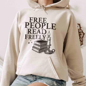 May include: Beige hoodie with the text "FREE PEOPLE READ FREELY" in black. Below the text is a graphic of a stack of books, a candle, and stars. The hoodie has a drawstring hood and a front pocket.