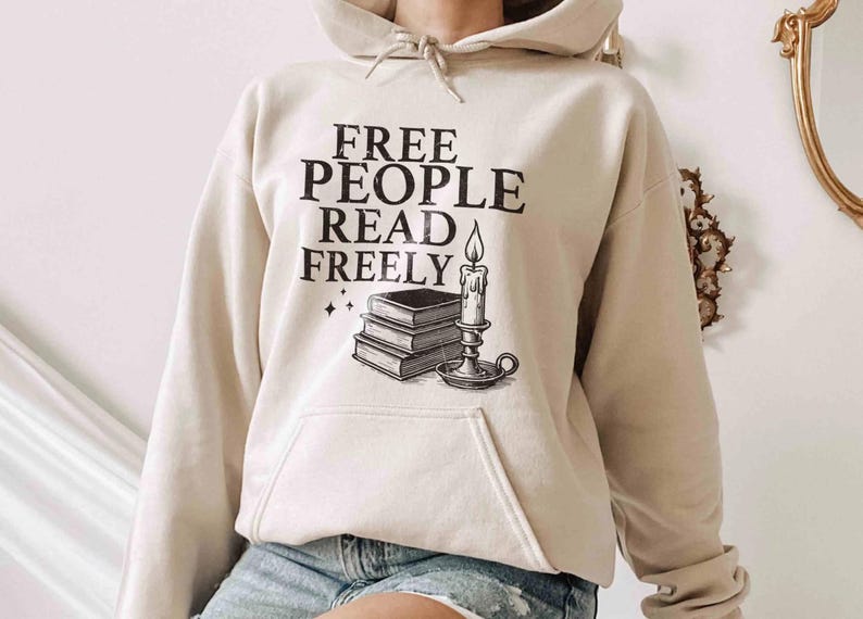 May include: Beige hoodie with the text "FREE PEOPLE READ FREELY" in black. Below the text is a graphic of a stack of books, a candle, and stars. The hoodie has a drawstring hood and a front pocket.
