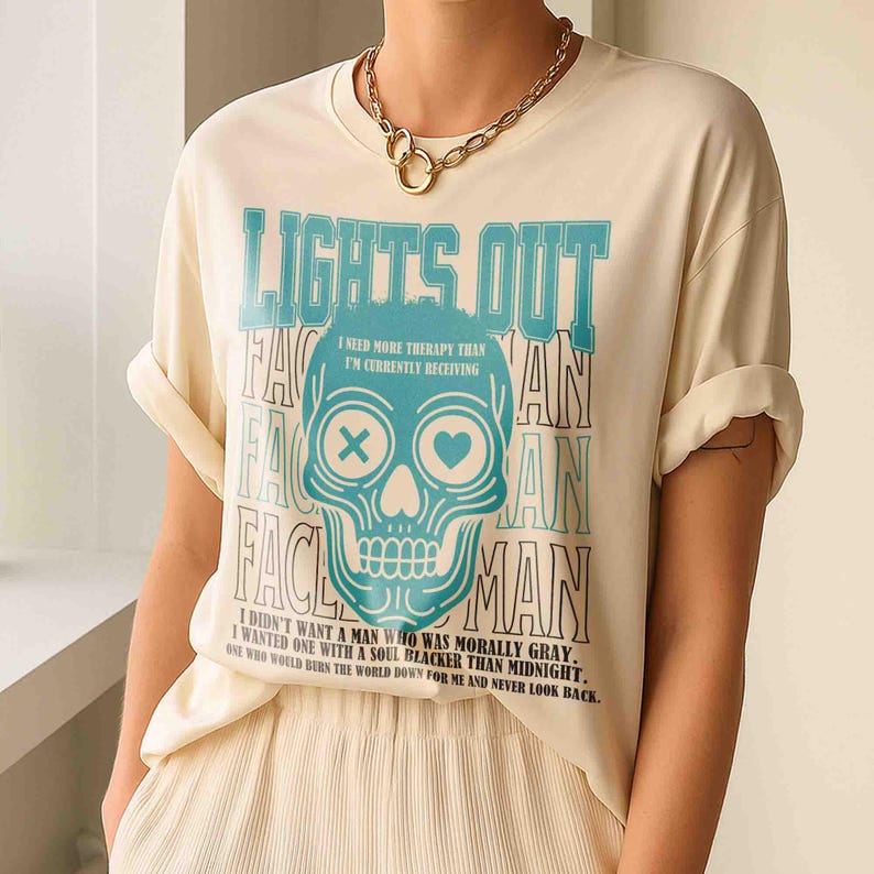 Pu&ograve; includere: T-shirt color crema con grafica a teschio turchese e le parole "LIGHTS OUT" e "VAIN". Il teschio ha una X per un occhio e un cuore per l'altro. La maglietta ha anche il testo "I need more therapy than I'm currently receiving."